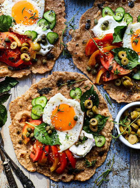 Mediterraner Frühstücksfladen mit Spiegelei, Tomaten, Gurken, Minos BIO Nativem Olivenöl Extra, Minos BIO Grünen Oliven mit Mandeln und Minos BIO Kalamata Oliven, serviert auf knusprigem Vollkornbrot.