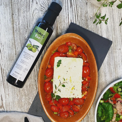 Eine Flasche Minos Bio Natives Olivenöl Extra neben einem Gericht mit frischen Kirschtomaten und Feta, das auf einem Holzbrett serviert wird, ideal für mediterrane Rezepte.