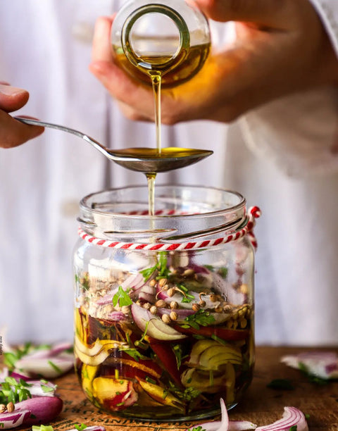 Ein Glas mit hausgemachtem Zwiebel-Kräuter-Dressing, zubereitet mit Minos Natives Olivenöl Extra, das aus einer Flasche auf einen Löffel gegossen wird – perfekt für frische Salate und Marinaden.