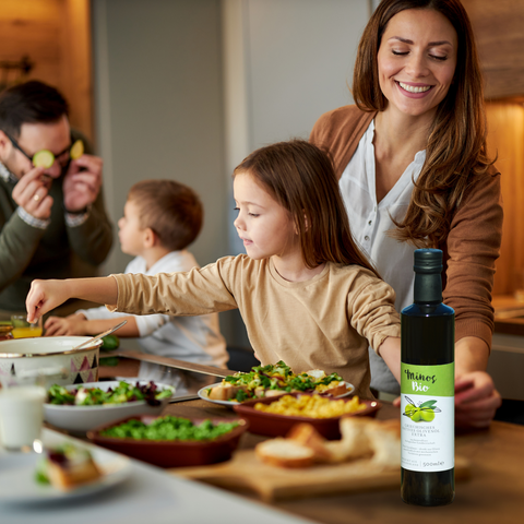 Familie bereitet zusammen eine Mahlzeit in der Küche vor, während sie Minos Bio-Olivenöl Extra verwenden. Die Kinder helfen, während die Mutter das Essen anrichtet, und das Olivenöl steht prominent auf dem Tisch, was auf seine Bedeutung für gesunde, familiäre Ernährung hinweist