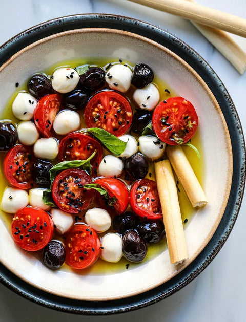 Frischer mediterraner Salat mit Kirschtomaten, schwarzen und grünen Oliven, Mozzarellakugeln und Grissini, verfeinert mit Minos Nativem Olivenöl Extra und garniert mit Basilikum und schwarzem Sesam.