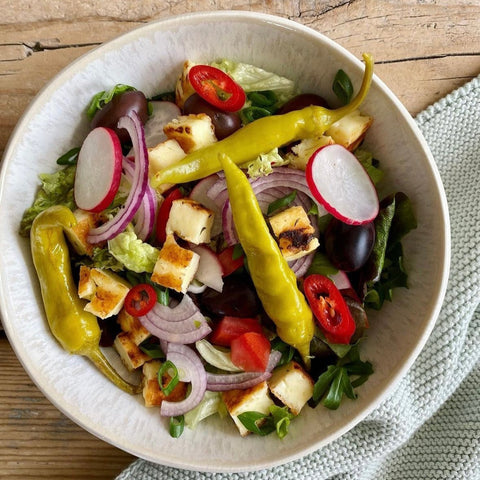 Frischer Salat mit knackigem Gemüse, roten Zwiebeln, Radieschen und Halloumi, garniert mit mild-pikanten Minos Pfefferoni und roten Chilis, serviert in einer weißen Schüssel auf einem Holztisch