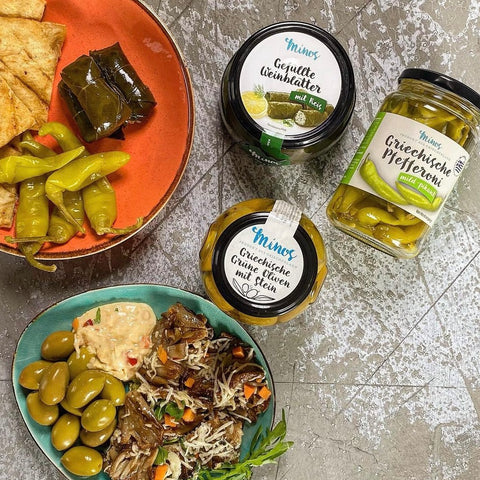  Ein mediterranes Gericht mit Minos Produkten: gefüllte Weinblätter, Grüne Oliven mit Stein, Griechische Pfefferoni mild-pikant und weitere mediterrane Zutaten, angerichtet auf farbenfrohen Tellern.