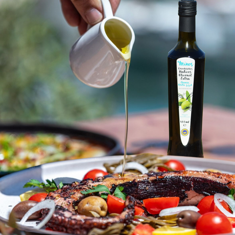Person gießt Minos natives Olivenöl Extra aus einer Flasche über eine mediterrane Gemüsepfanne, die mit frischen Zutaten und Oliven garniert ist.